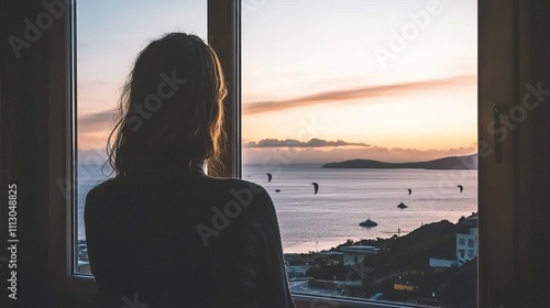 A contemplative woman gazes out a window at a stunning sunset over the ocean.