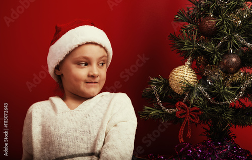 Emotional kid girl with surprising eyes looking on the decor holiday fur green tree with gift in red santa claus hat. Happy expression winter holadays with natural emotion child photo