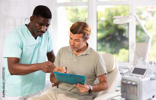 Interested middle aged male patient signing informed consent form while sitting on couch during consultation with cosmetologist. Concept of paperwork when receiving personalized medical services