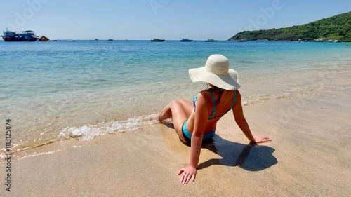 Young woman wearing a sun hat sitting on the sand of a tropical beach enjoying summer vacation