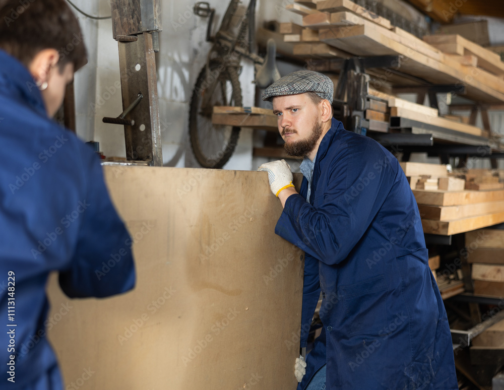 Two workers at a furniture factory move a sheet of plywood onto a planing machine together Two workers at a furniture factory move a sheet of plywood onto a planing machine together