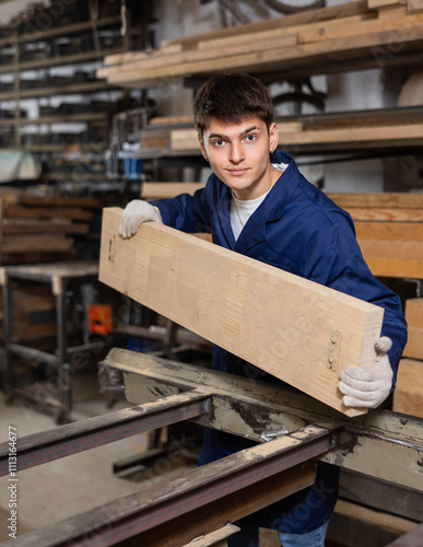 Young male carpenter putting wooden board on cutting machine in wood workshop