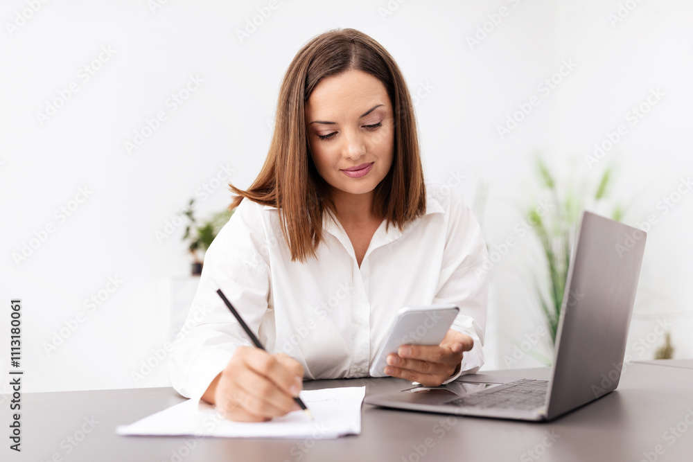 A businesswoman actively engages in her work at a modern office. She uses a smartphone to check information while taking notes on a notepad, immersed in her tasks. A businesswoman actively engages in her work at a modern office. She uses a smartphone to check information while taking notes on a notepad, immersed in her tasks.