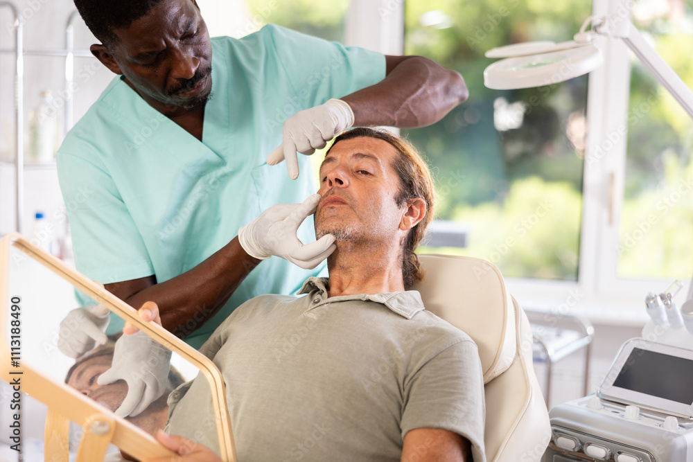 Fototapeta premium Adult man looking in mirror with interest while skilled African American male cosmetologist showing results of hardware facial procedure in office of aesthetic medicine clinic