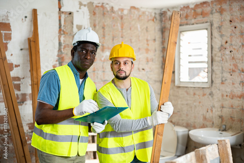 Portrait of two builders, african-american with documents and caucasian man with wooden plank, standing inside construction site. photo