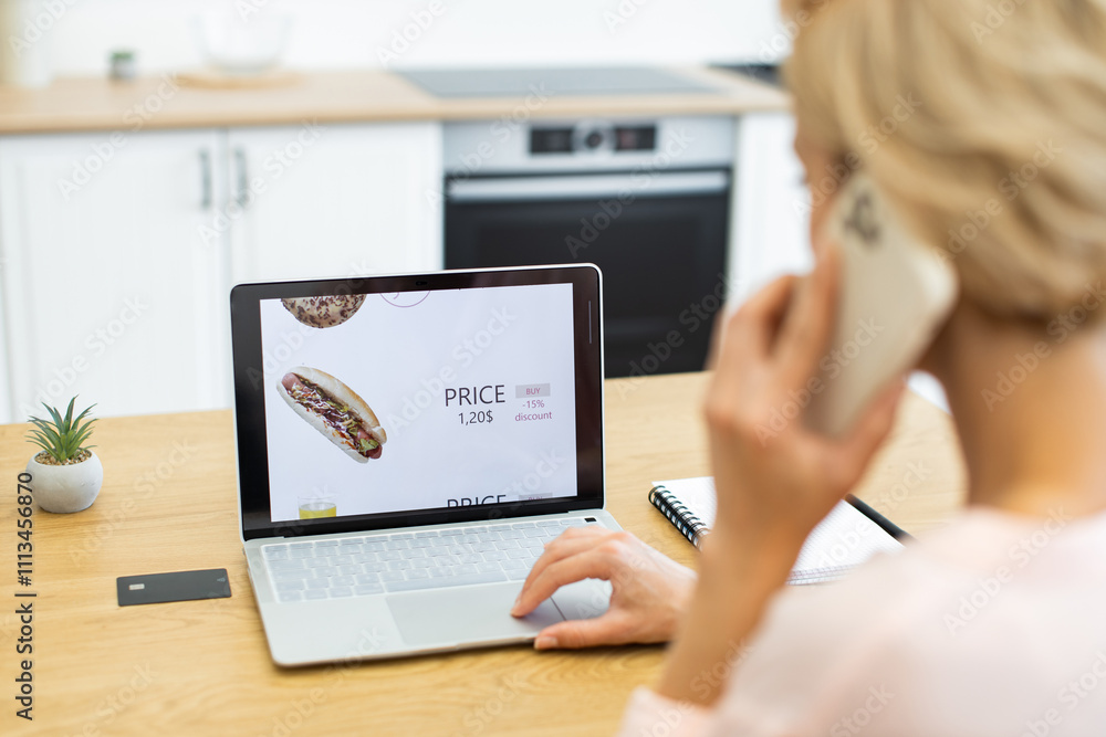 Caucasian woman browsing online food delivery website on laptop while talking on phone. Modern kitchen setting with potted plant on wooden table. Scene emphasizes convenience and technology. Caucasian woman browsing online food delivery website on laptop while talking on phone. Modern kitchen setting with potted plant on wooden table. Scene emphasizes convenience and technology.