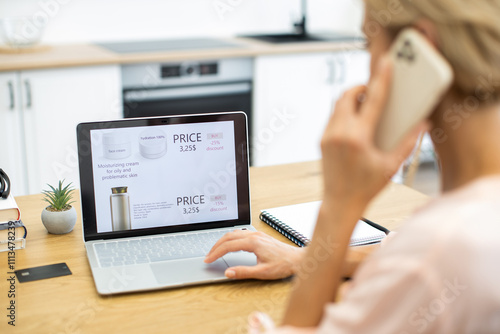 Caucasian woman multitasking with phone and laptop while ordering cosmetics online. She is browsing an ecommerce website, focused on skincare products, displaying discounted prices.