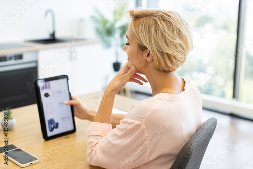 Caucasian woman in 30s shopping online using tablet at home. She is viewing clothing options on delivery website while sitting casually in a modern, bright kitchen.