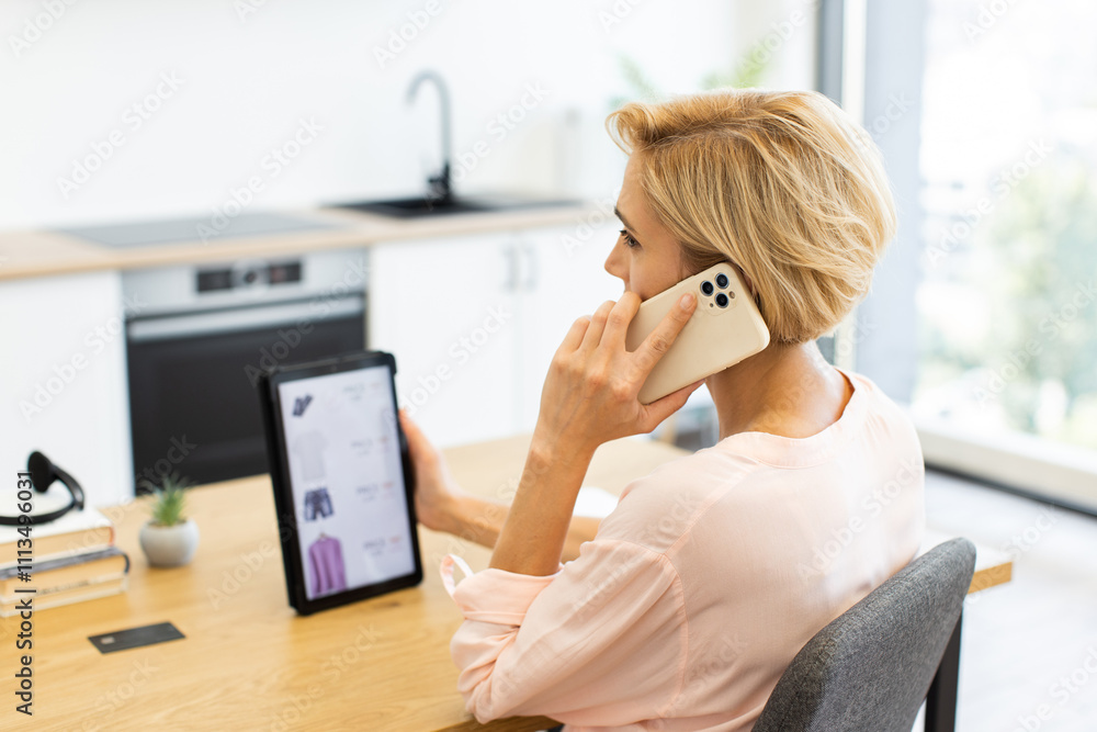 Obraz premium Caucasian woman using smartphone while browsing clothing website on tablet. Sitting at desk in modern kitchen, ordering clothes online with delivery service.
