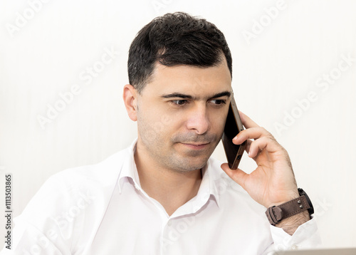 man working in office,talking at smartphone or having video call.guy with headphones is smiling,explain something,gesturing with hands. worker writing something in agenda.monstera leaf in background.