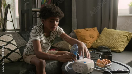 Little Black boy sitting on sofa at home, playing video game on console, then coughing and inhaling liquid medicine with nebulizer to treat asthma