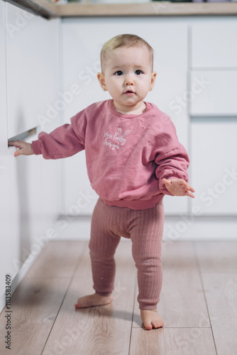 a one-and-a-half-year-old girl stands against the background of the kitchen