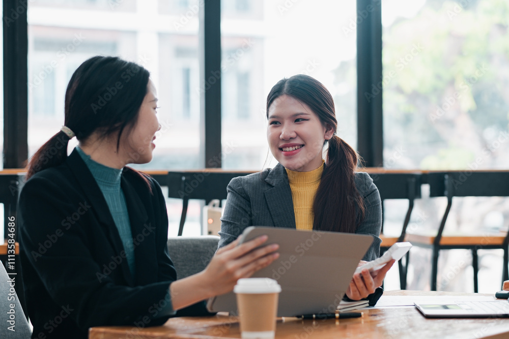 Two women in business attire discuss work while using a tablet and smartphone in a contemporary office environment. Two women in business attire discuss work while using a tablet and smartphone in a contemporary office environment.