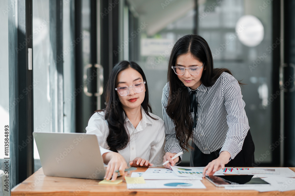 Two women working together in a modern office, reviewing documents and using a laptop for a business project. Two women working together in a modern office, reviewing documents and using a laptop for a business project.