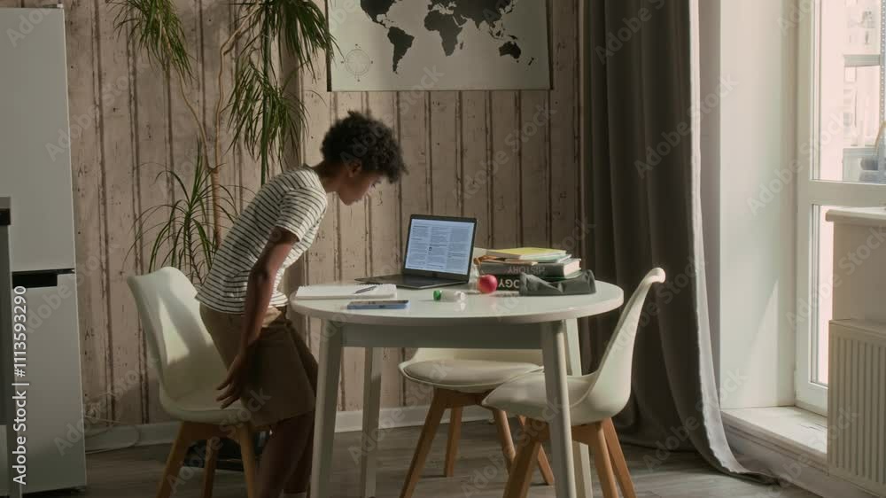 Young African American boy walking in kitchen, sitting at table with books and asthma inhaler on it, reading text on laptop screen and writing down notes on paper while doing school homework Young African American boy walking in kitchen, sitting at table with books and asthma inhaler on it, reading text on laptop screen and writing down notes on paper while doing school homework