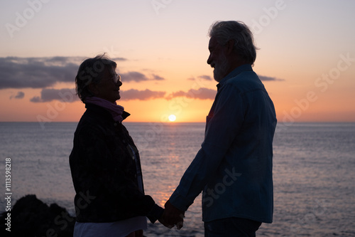 Silhouette of senior couple standing on the rocks at sea looking each other in the eyes while the sun goes down. Relaxed caucasian man and woman enjoying vacation or retirement photo