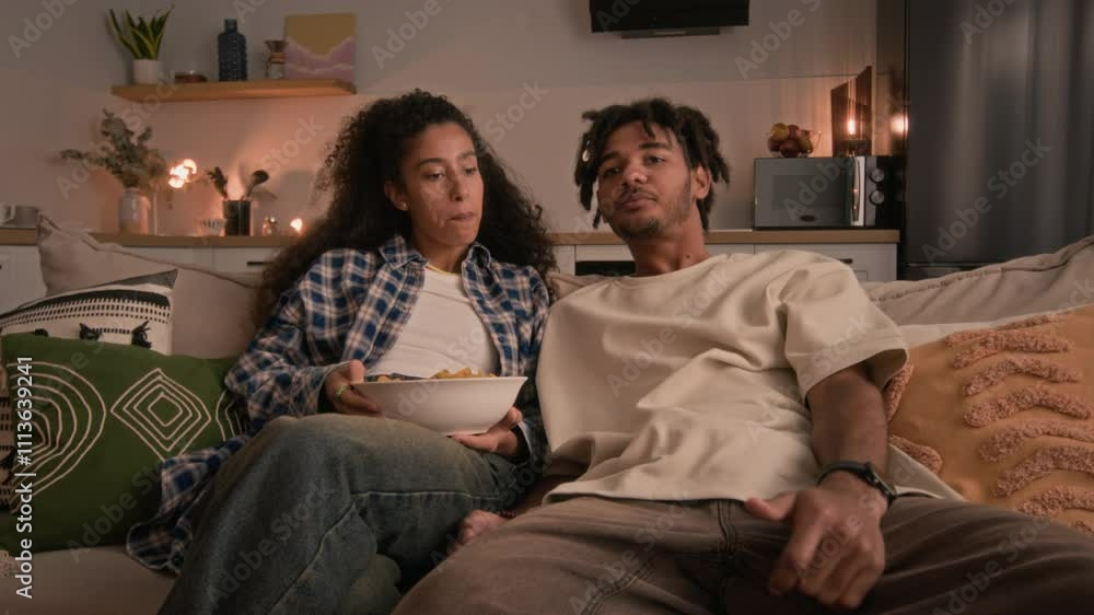 Medium shot of Biracial young couple in casualwear sitting on couch in bright cozy living room setting, sharing snacks and chatting while relaxing in front of TV together at weekend Medium shot of Biracial young couple in casualwear sitting on couch in bright cozy living room setting, sharing snacks and chatting while relaxing in front of TV together at weekend