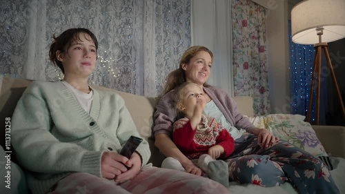 A family enjoy quality time together in the evening, watching TV in their cozy living room. Mother and children sitting comfortably on a couch, surrounded by warm lighting and a relaxed atmosphere
