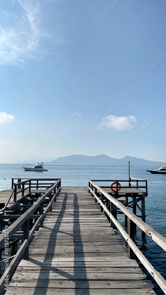 Obraz premium Rustic dockyard wooden deck footpath on the open water, with beautiful mountain background, vast blue sea, under a clear sunny blue sky. Beautiful landscape and holiday feel.