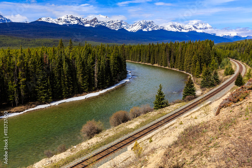 Canvas Print A train tracks run along a river with mountains in the background