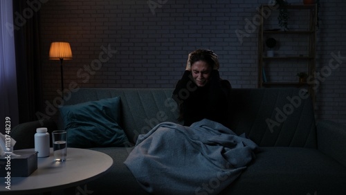 Woman suffering mental health issues, sitting on the sofa covered in blanket, depressed expression, tense emotions, holding head.