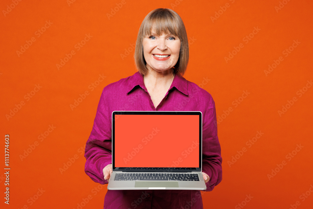 Elderly blonde woman 50s years old she wear purple shirt casual clothes hold use work on laptop pc computer with blank screen isolated on plain red orange background studio portrait Lifestyle concept Elderly blonde woman 50s years old she wear purple shirt casual clothes hold use work on laptop pc computer with blank screen isolated on plain red orange background studio portrait Lifestyle concept