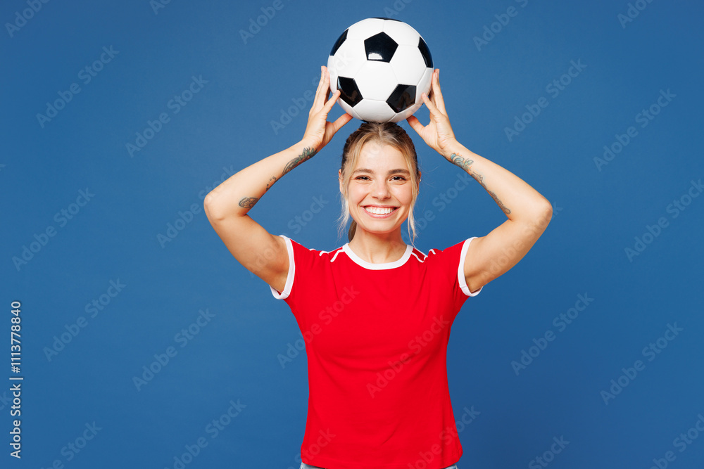 Young smiling cheerful happy woman fan wear basic red t-shirt cheer up support football sport team hold in hand soccer ball watch on head tv live stream look camera isolated on plain blue background.