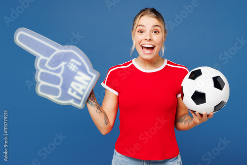 Young happy woman fan in basic red t-shirt foam 1 fan glove finger up point aside cheer up support football sport team hold in hand soccer ball watch tv live stream isolated on plain blue background photo