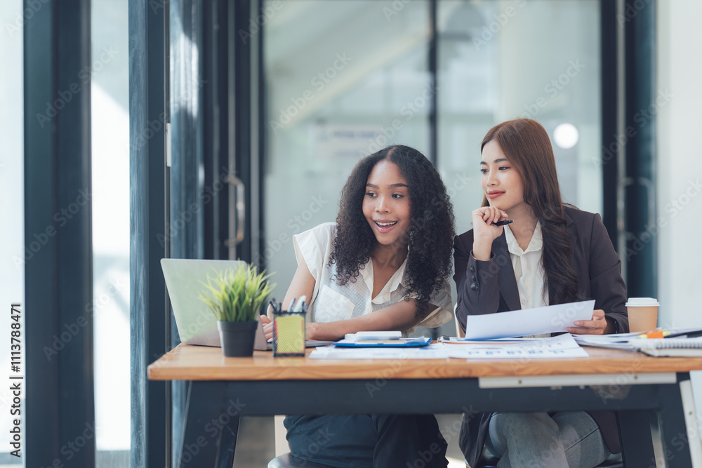 Obraz premium Collaborative Brainstorming: A diverse team of two young businesswomen strategize and discuss ideas, leaning in over a laptop and documents.