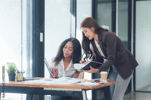 Collaborative Brainstorming: Two businesswomen, one Asian and one Black, are working together on a project. They are reviewing a document on a tablet.