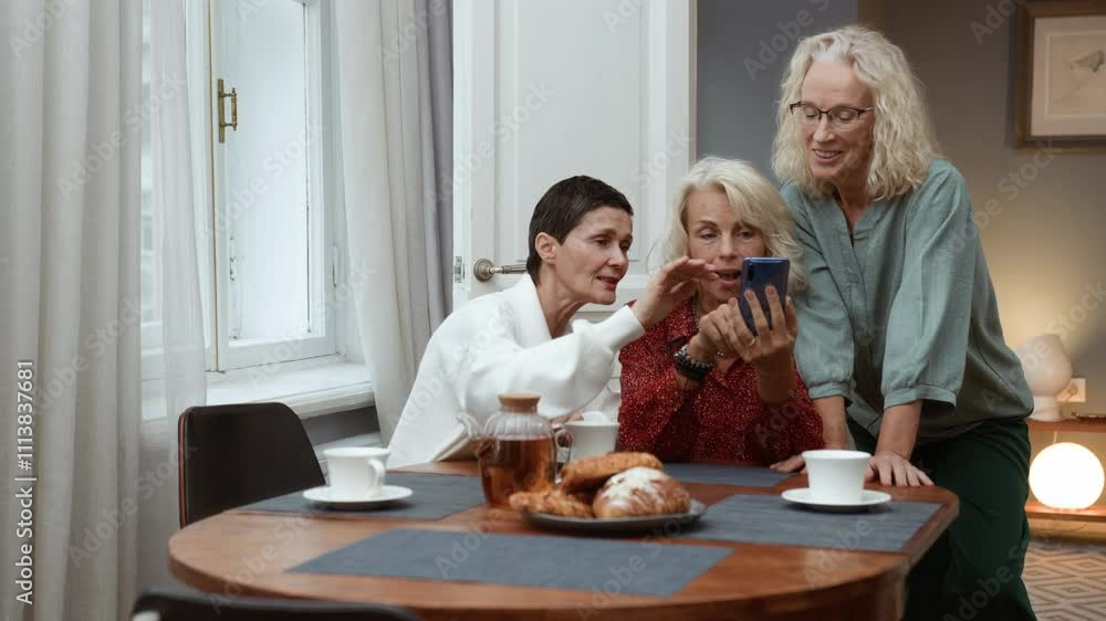 Three elderly women sit at table chatting joyfully, looking in mobile telephone. Elegance blonde senior women chill together talking. Female aged friends have tea time at home and communicate smiling Three elderly women sit at table chatting joyfully, looking in mobile telephone. Elegance blonde senior women chill together talking. Female aged friends have tea time at home and communicate smiling