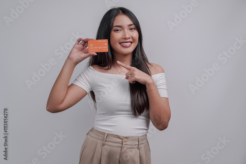 Cheerful young Asian woman pointing to the credit card she holds, isolated by white background.