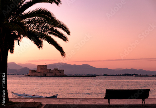 Postcard framed photo of beautiful and romantic walkway in Nafplio with Bourtzi fortress in the background and sun setting in golden and purple tones over the mountains.
