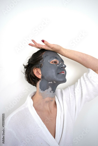 Woman in a white robe with a gray facial mask looking up with her hand on her head against a white background. photo