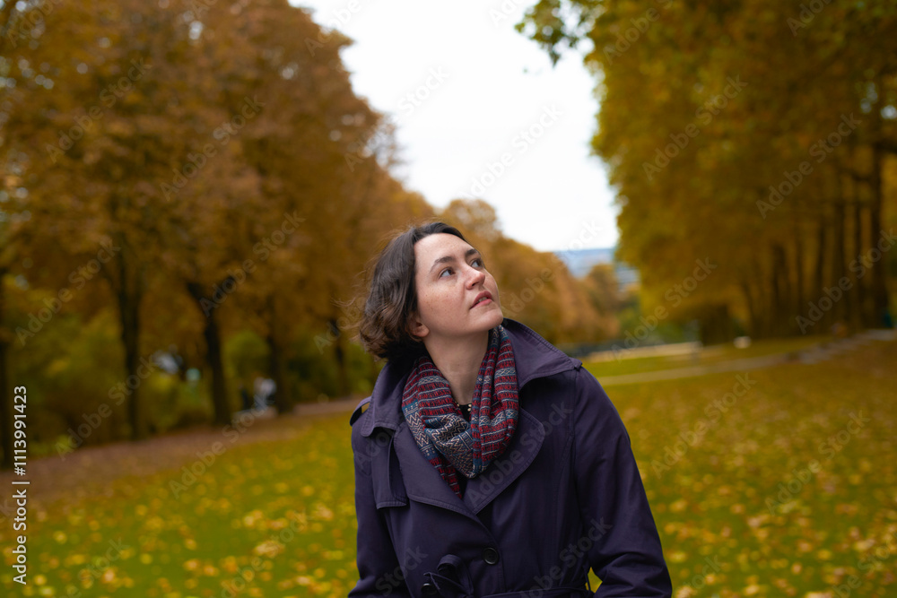 Woman in a navy coat looking up while standing in a park with autumn trees and fallen leaves. Woman in a navy coat looking up while standing in a park with autumn trees and fallen leaves.