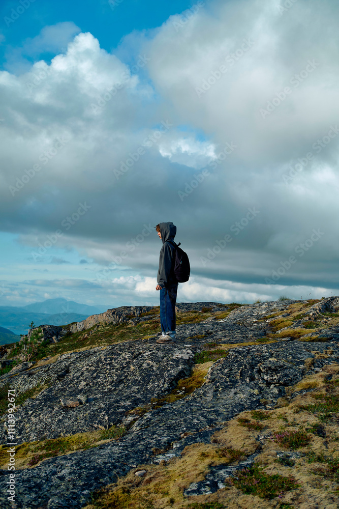 A person in a hoodie and backpack stands on a rocky hilltop overlooking a sweeping landscape under a cloudy sky. A person in a hoodie and backpack stands on a rocky hilltop overlooking a sweeping landscape under a cloudy sky.