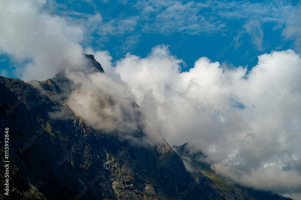 Clouds shroud the rugged peaks of a mountain under a bright blue sky, Trollstiege, Trollvegen, Norway Clouds shroud the rugged peaks of a mountain under a bright blue sky, Trollstiege, Trollvegen, Norway