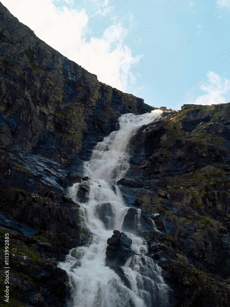 A cascading waterfall flows down a rugged cliff face against a bright blue sky backdrop, Trollstiege, Trollvegen, Norway A cascading waterfall flows down a rugged cliff face against a bright blue sky backdrop, Trollstiege, Trollvegen, Norway