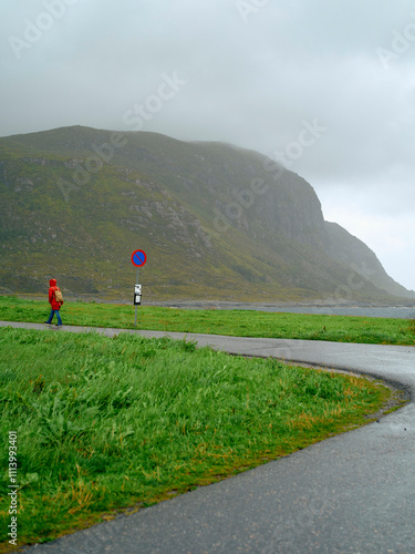 Solo hiker in red walking beside a rural road with a no parking sign and a mountainous background under overcast skies, Norway photo