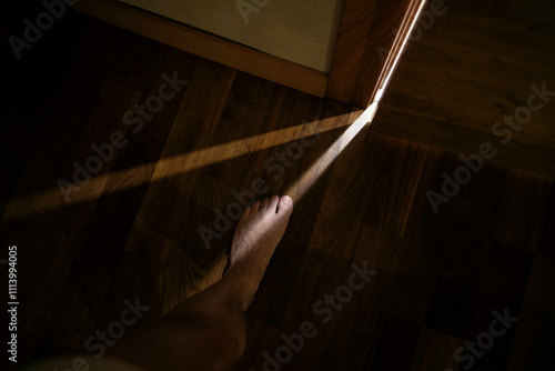 Barefoot person standing on a wooden floor with a beam of sunlight casting shadows. photo