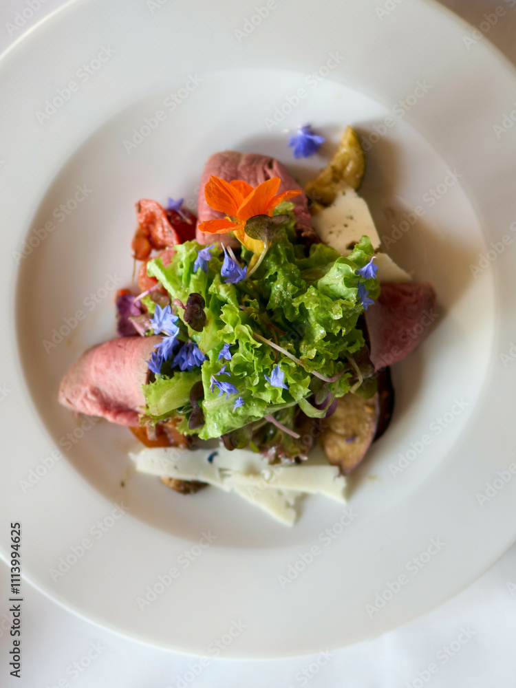 A gourmet salad served during wedding on a white plate with fresh greens, edible flowers, roasted vegetables, and sliced meat. A gourmet salad served during wedding on a white plate with fresh greens, edible flowers, roasted vegetables, and sliced meat.
