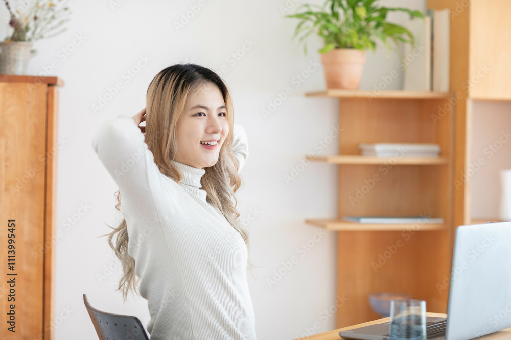 Smiling woman with long wavy hair sitting at a desk with a laptop, stretching with hands behind her head in a brightly lit room. Smiling woman with long wavy hair sitting at a desk with a laptop, stretching with hands behind her head in a brightly lit room.