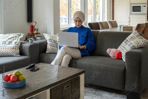 A woman in a hijab sits on a grey couch working on a laptop in a cozy living room with a bowl of fruit on the coffee table. photo