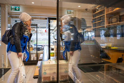 Woman in a white headscarf and jeans looking at items in a refrigerated store display with her reflection visible on the glass, Netherlands photo