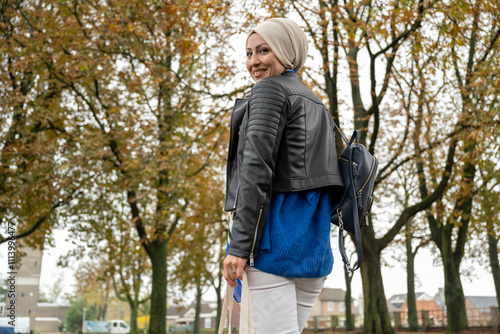 A smiling woman in a hijab and leather jacket carries a backpack and looks back over her shoulder in an urban setting with autumn trees, Netherlands photo