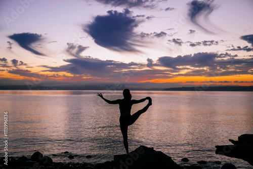 Silhouette of a woman practicing yoga on a rocky shoreline at sunset with orange and purple hues in the sky. photo