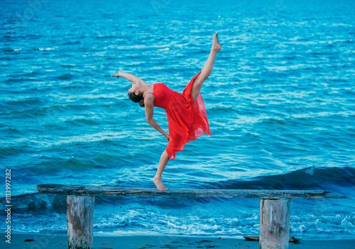 A woman performs an elegant dance move on a wooden beam by the sea, wearing a flowing red dress, USA photo