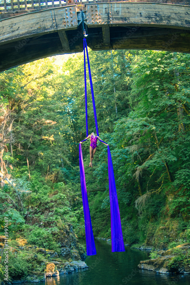 © Connect Images - An aerial performer demonstrates silk acrobatics beneath a wooden bridge surrounded by lush forest, USA