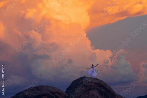 A woman in a white dress stands triumphant atop a rock formation under a vibrant sunset sky, USA
