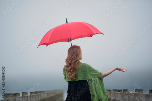 Redheaded woman holding a bright red umbrella stands on a foggy pier reaching out her hand to feel the mist. photo